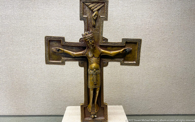 Medieval metal crucifix with elongated Christ figure on a dark cross with squared terminals and upper plaque.