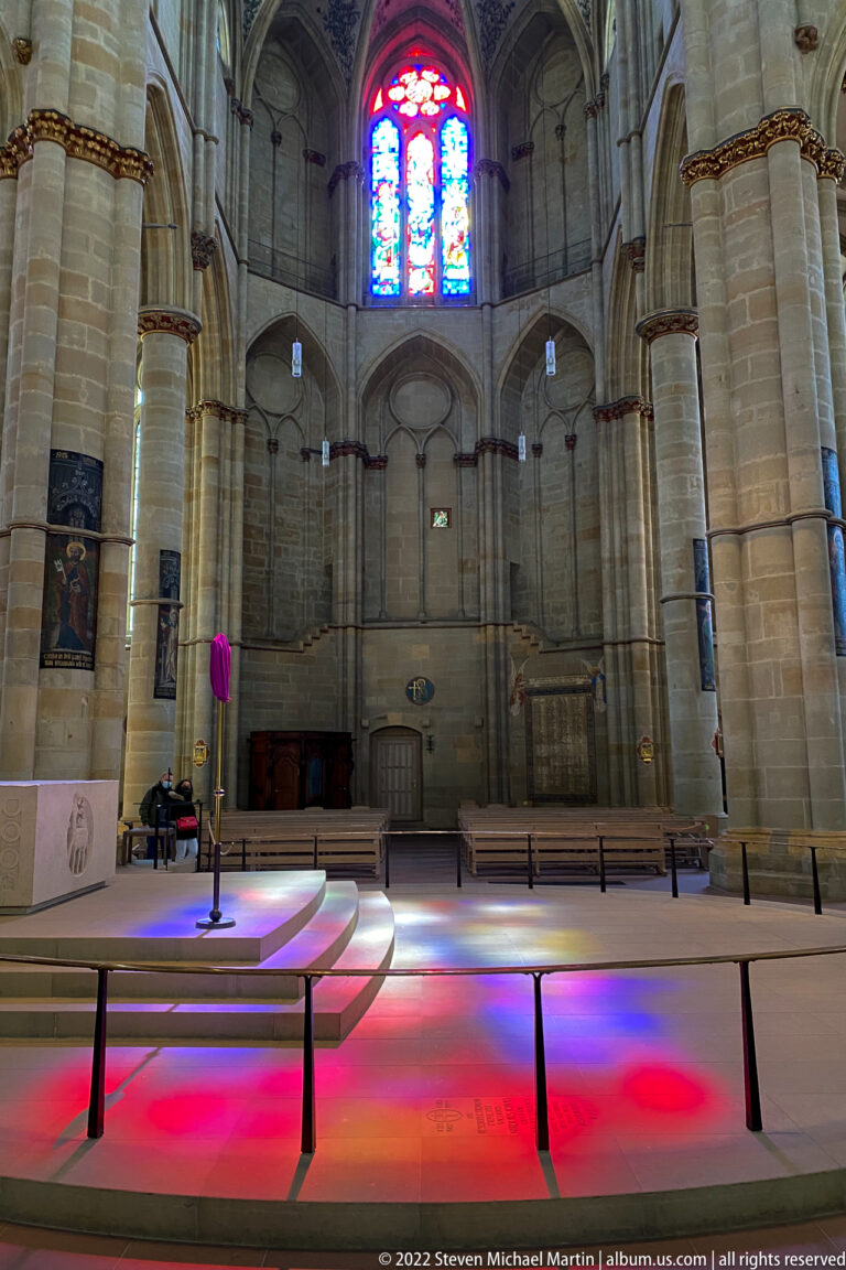 Gothic church interior with tall columns, pointed arches, stained-glass window, and vivid colored light across the floor.