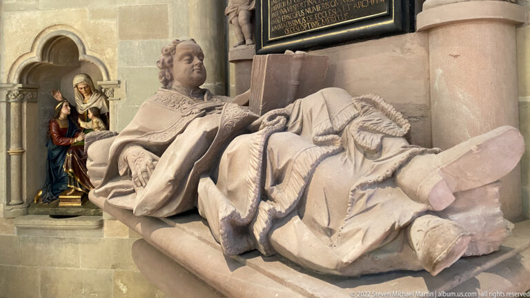 Reclining stone effigy of a robed cleric holding an open book, with a devotional niche sculpture behind.