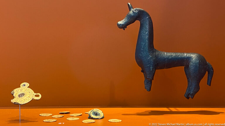 Dark bronze horse figurine displayed with coins and small metal ornaments against an orange museum background.