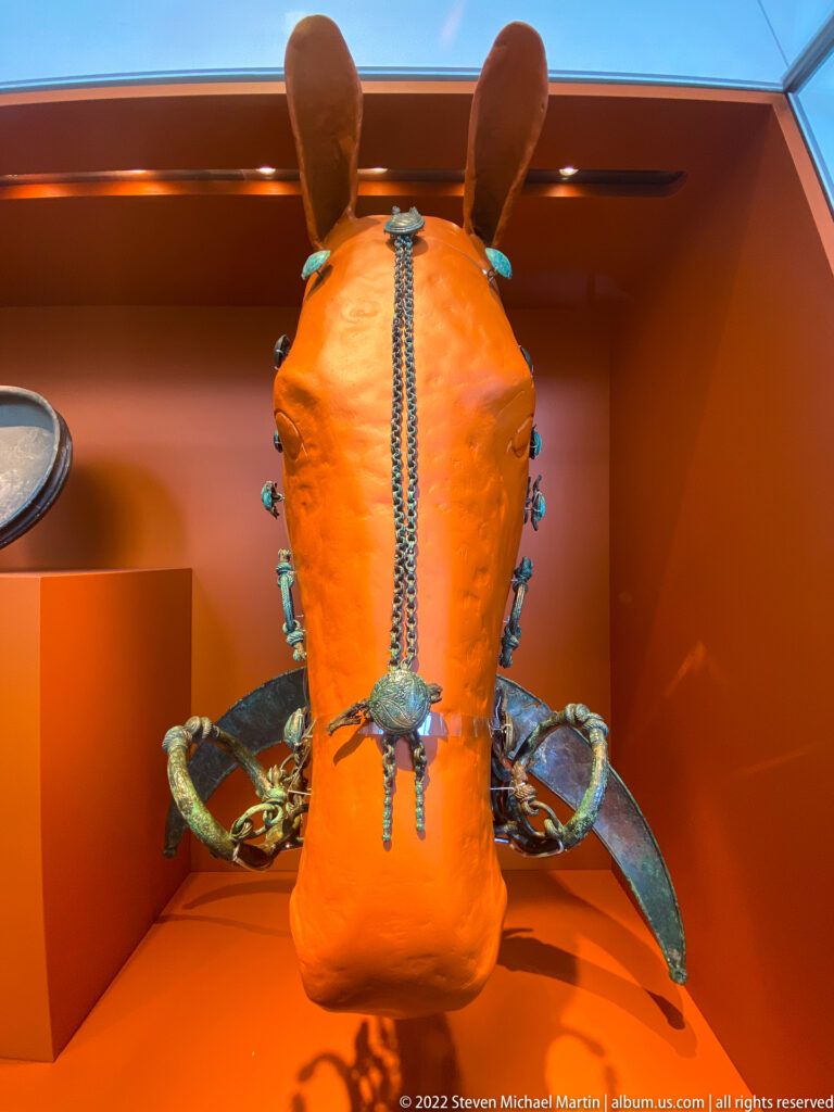 Reconstructed horse tack with chains, pendants, green fittings, and broad metal pieces mounted on an orange horse-shaped support.