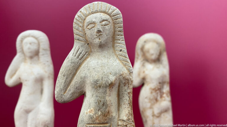 Small pale standing female figurine with stylized face and radiating hair, flanked by two similar blurred figures.