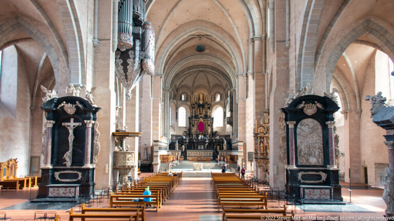 Central aisle of Trier Cathedral with wooden seating, stone piers, ribbed vaults, side monuments, and high altar beyond.