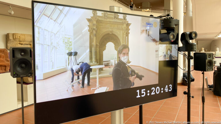 Interactive museum installation with live video screen, visitor, cameras, speakers, and a digital time display.