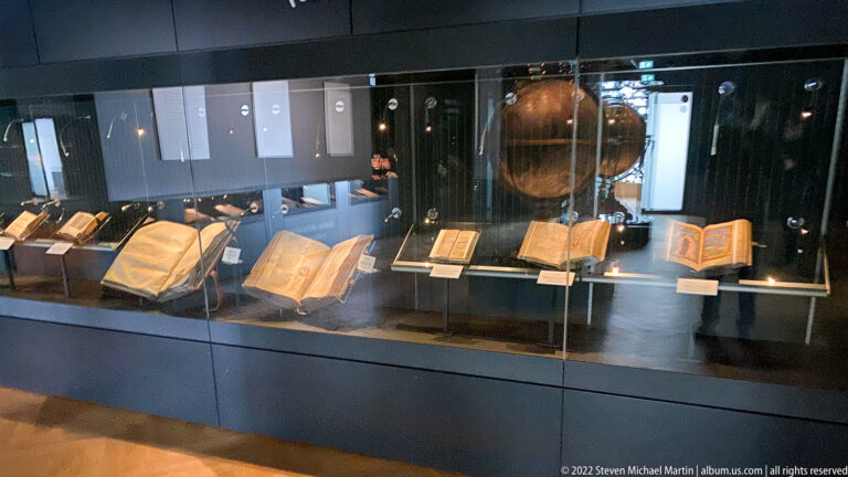 Glass museum case with several open historic books and manuscripts arranged in a row under dark gallery lighting.