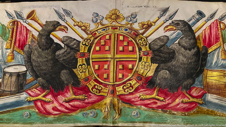 Red-and-gold coat of arms supported by two black ravens, with flags, spears, drums, and military trophies behind.