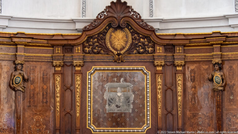 Dark carved wooden cabinet or shrine with gilded trim, central glazed panel, and radiant emblem above.