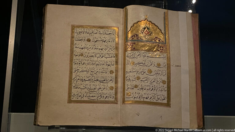Open Arabic-script manuscript with black calligraphy, gold details, and an illuminated gold-and-blue heading panel.