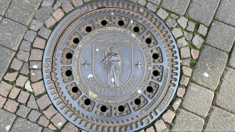 Round metal manhole cover in stone paving marked STADT TRIER around a central civic emblem.