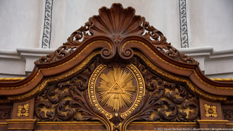 Gilded Eye of Providence within a radiant oval, surrounded by dark carved scrollwork and a shell-shaped crest.