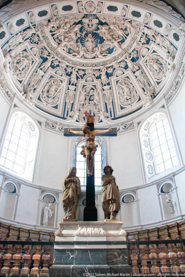 Crucifix and sculpted figures on a dark marble base beneath a bright, richly stuccoed domed ceiling.