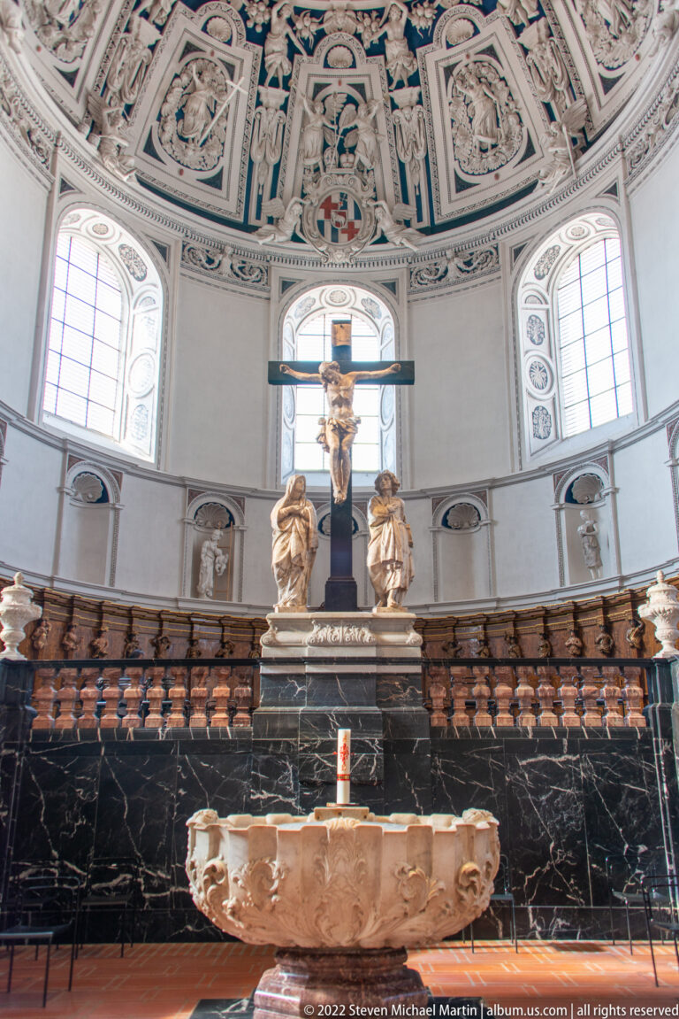 Stone altar in front of a crucifix and sculpted figures, with curved choir seating and bright chapel windows behind.