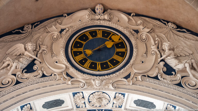 Black clock with gold Roman numerals set in ornate white stucco with winged figures and scrolling decoration.
