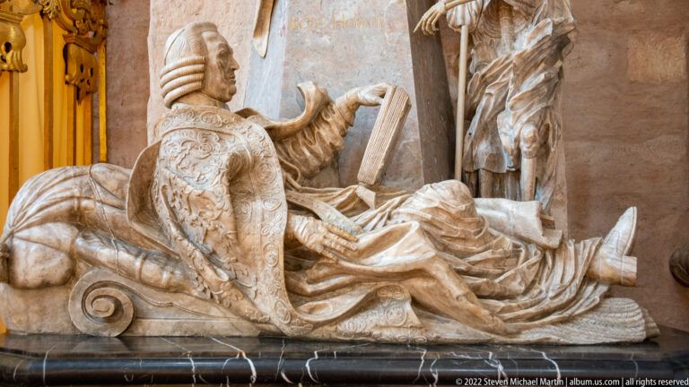 Reclining armored stone figure on a scroll-ended support beside drapery and part of a standing figure.