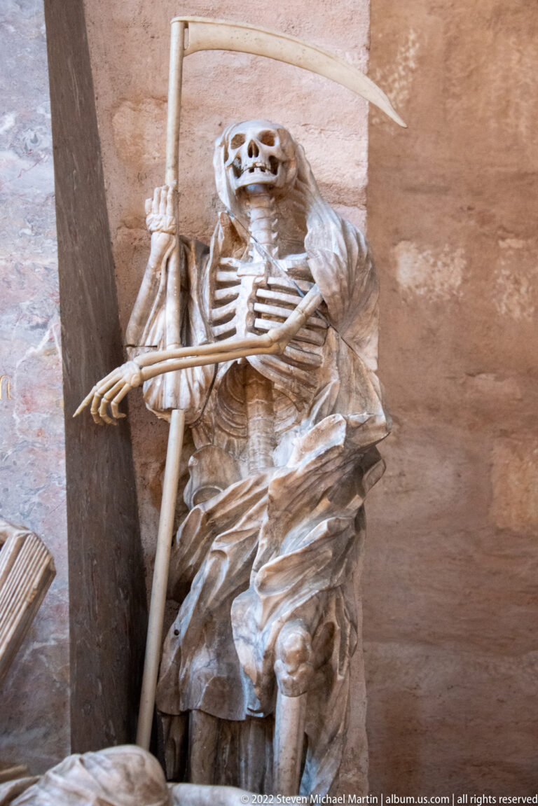 Carved skeleton seated with a long scythe and draped cloth against a stone interior wall.