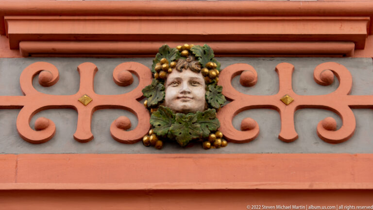 Sculpted face surrounded by green leaves and gold fruit on a terracotta-and-gray decorative façade.