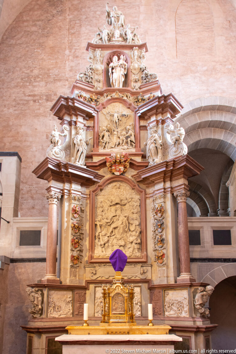 Tall Baroque altar with colored columns, sculpted figures, relief panels, gilded fittings, and a purple-covered central object.