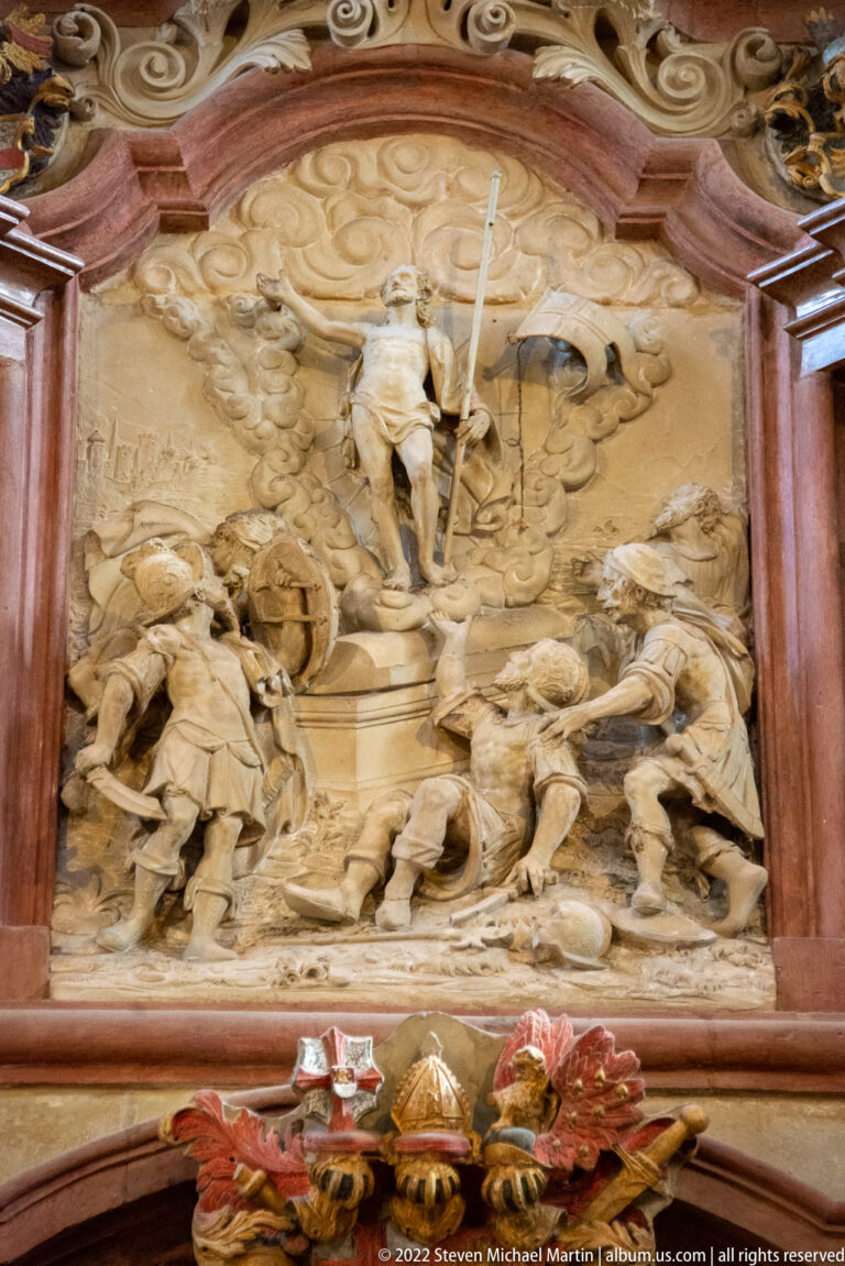 Pale carved religious relief with standing and kneeling figures, clouds, and flowing drapery inside a reddish altar frame.