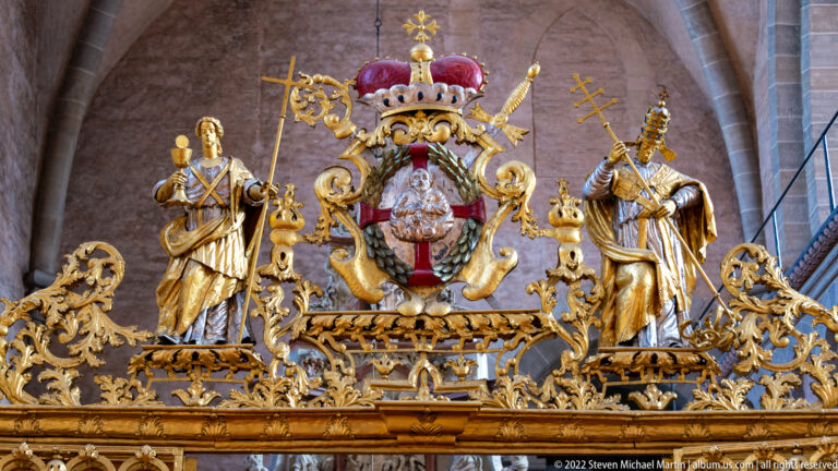 Large gilded crowned coat of arms flanked by two standing figures above ornate scrolling metalwork.