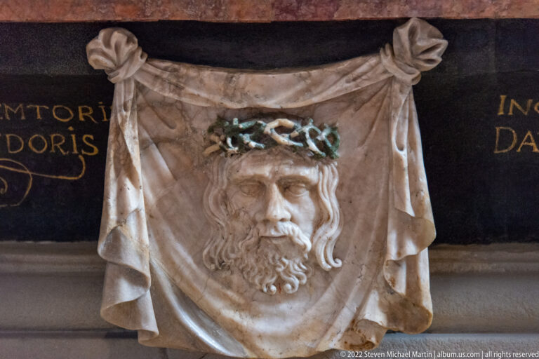 Relief of Christ’s bearded face on a draped, knotted cloth against a dark background with partial gold lettering.