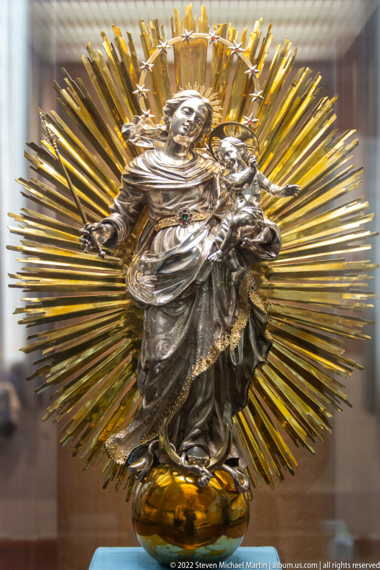 Silver Virgin and Child figure centered before a large gilded sunburst in a display case.
