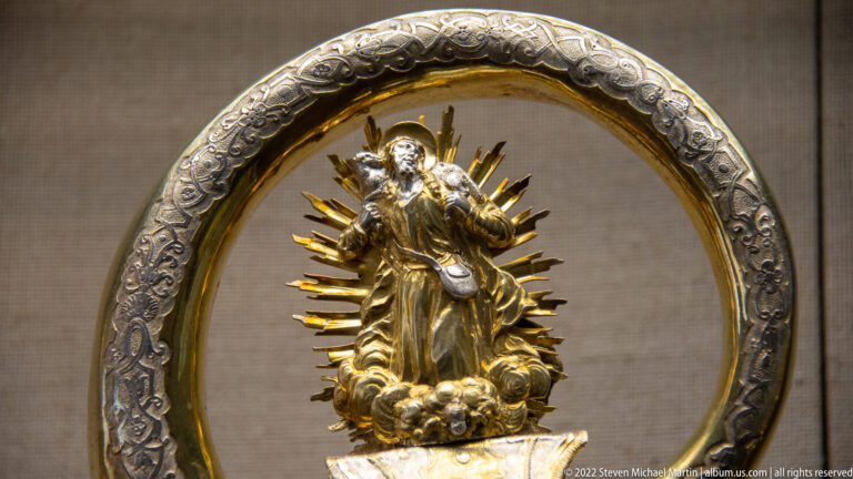 Small gilded female sacred figure standing on clouds with radiating rays inside an ornate oval frame.