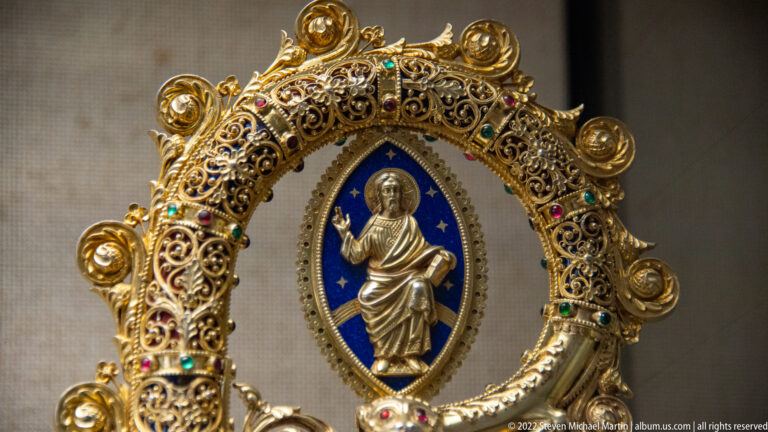 Standing Christ figure on blue enamel inside an ornate gold filigree frame set with colored gemstones and beads.