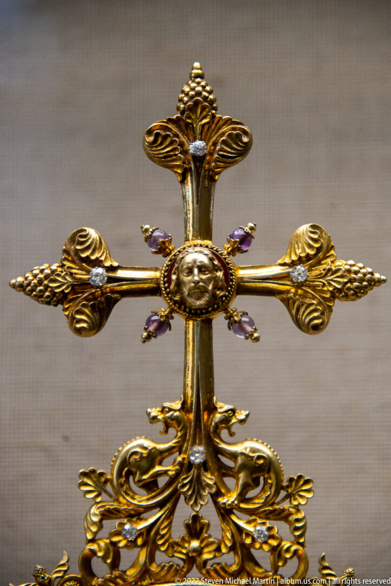 Ornate gilded cross with lobed ends, central round medallion, decorative base, and small purple jewel-like accents.