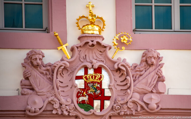 Crowned heraldic shield with sculpted supporters, crossed regalia, and scrollwork on a pink-and-white façade.