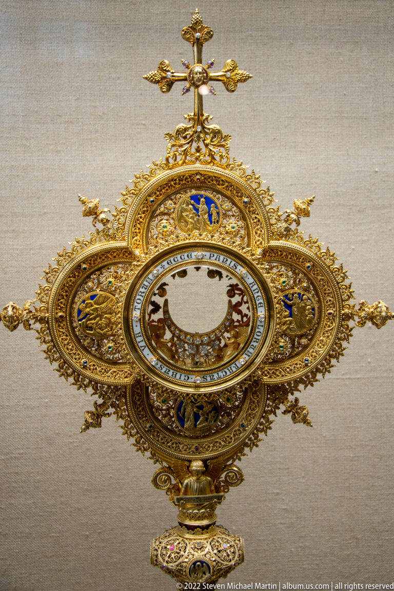 Circular gilded treasury object with white center, blue enamel, radiating filigree, lower stem, and small cross above.