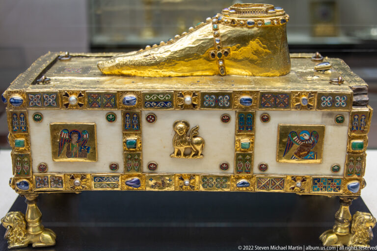 Gold foot-shaped reliquary above a rectangular enamelled casket with gemstones, miniature sacred images, and short supports.