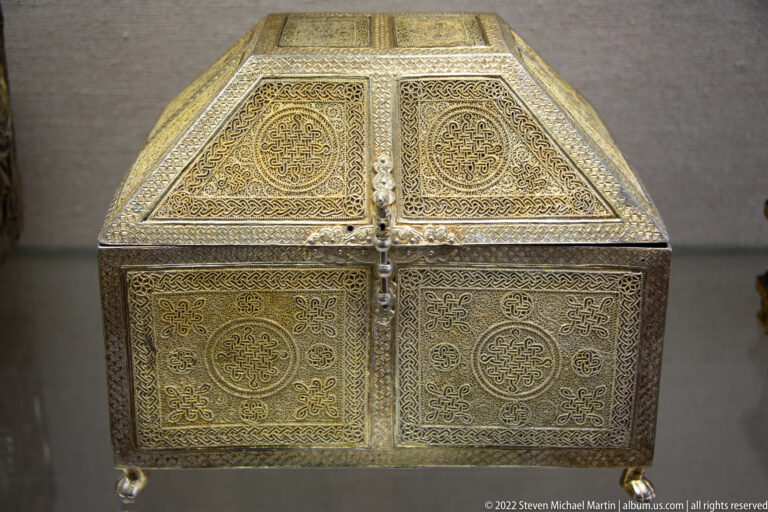 Small metal reliquary chest with a sloping lid, engraved geometric and foliate panels, and short feet.
