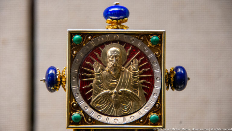 Square jeweled devotional object with a central silver relief of Christ, red backing, turquoise stones, and blue finials.