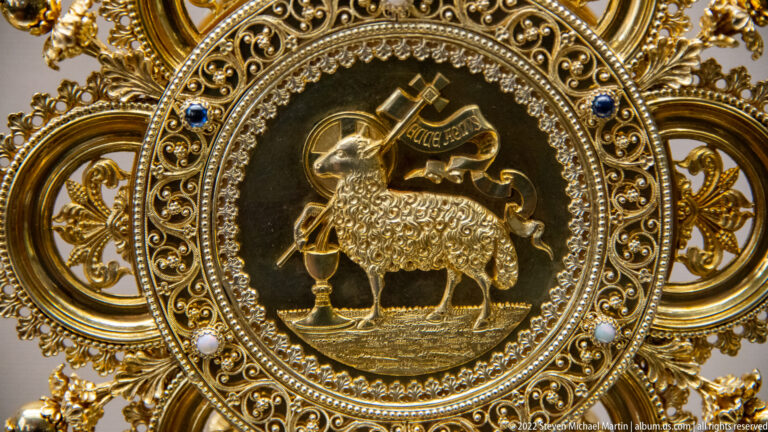 Gilded circular Agnus Dei medallion with lamb, cross-staff, banner, pearls, blue stones, and ornate scrollwork.