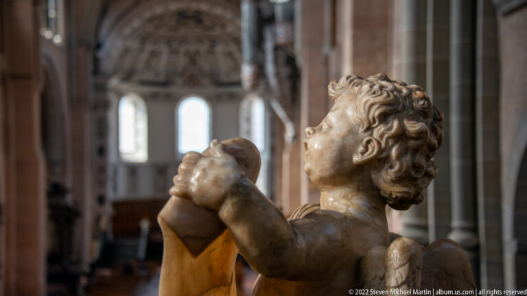 Carved child figure with clasped hands and upward gaze, seen against the blurred interior of Trier Cathedral.
