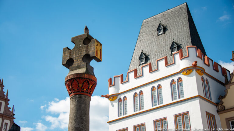Dark stone cross column in front of a white historic building with steep slate roof and red-trimmed stepped gable.