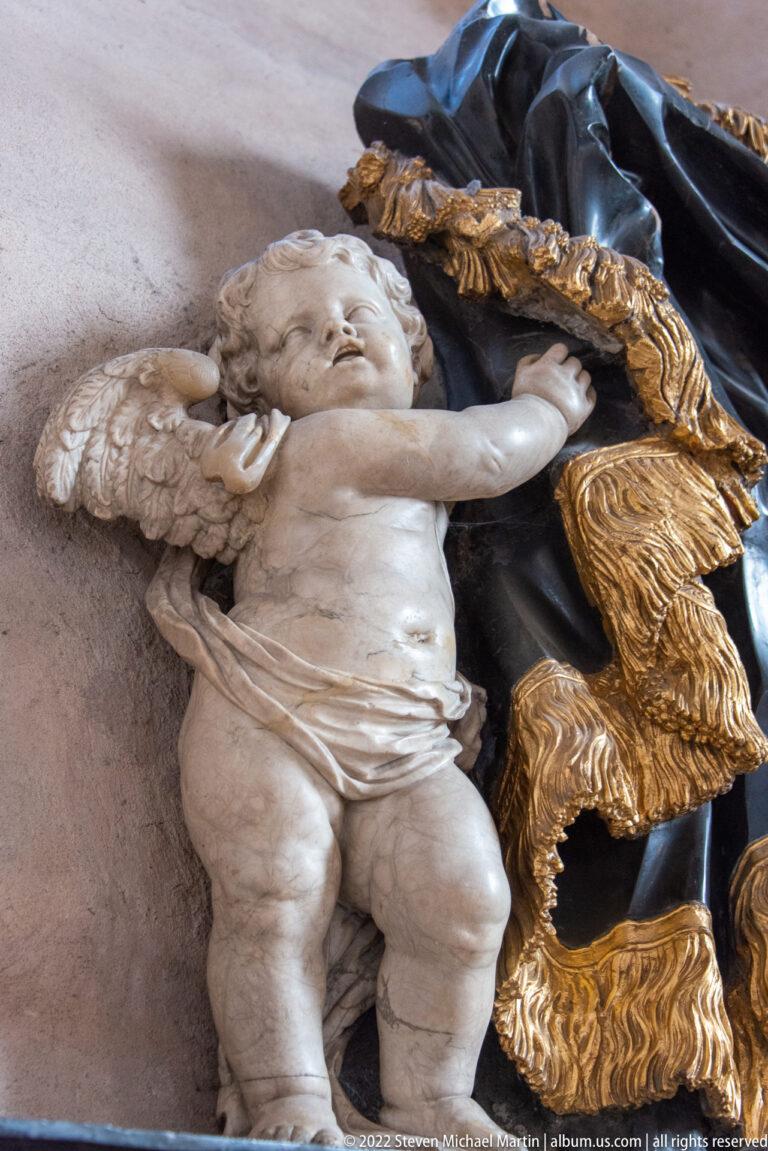 Reclining sculpted infant beside dark drapery with a thick carved fringe on a cathedral monument.