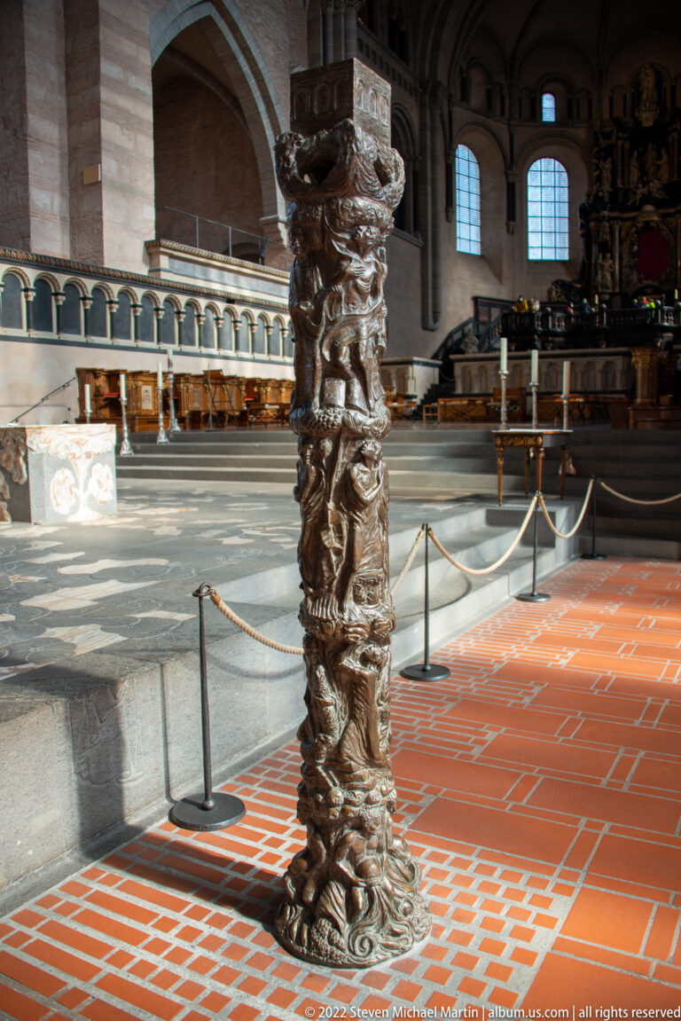 Tall dark ceremonial column or candlestick with dense figurative carving standing on the red-tiled cathedral floor.