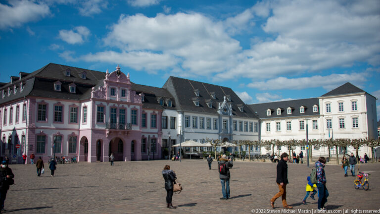 Cobbled Trier market square with pedestrians and a row of historic pink, white, and dark-roofed town houses.