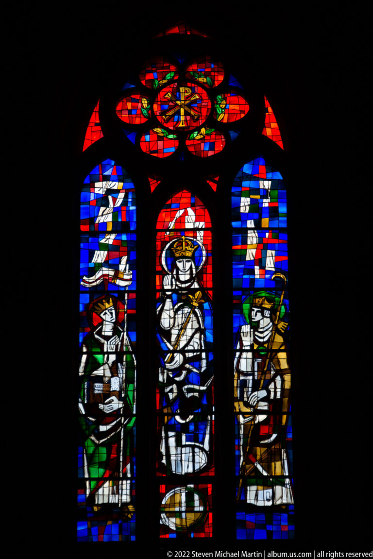 Three-lancet stained-glass window with a circular upper light and vivid red, blue, green, and white sacred figures.