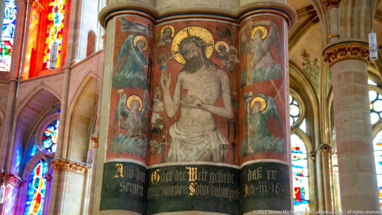 Painted Christ with haloed figures on a church pier above a black band of Gothic-script inscription.