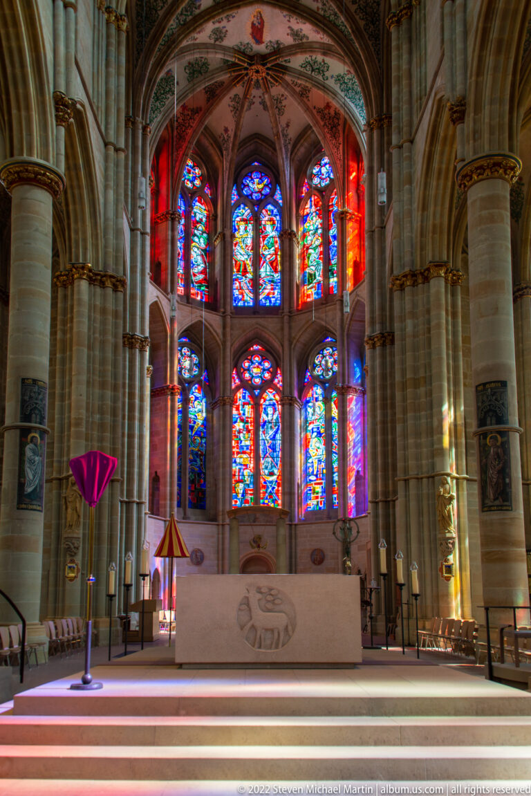 Modern altar before tall stained-glass windows and Gothic stone columns in a bright church sanctuary.