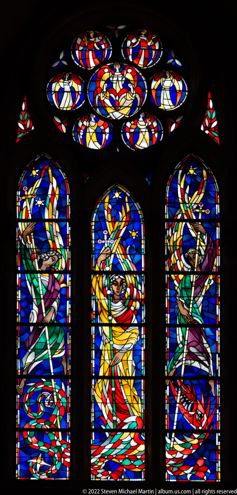 Three modern stained-glass lancets with stylized sacred figures in blue, yellow, green, and red beneath circular upper lights.