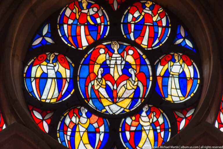 Circular stained-glass rose window with a central sacred scene surrounded by smaller red, blue, yellow, and white figures.