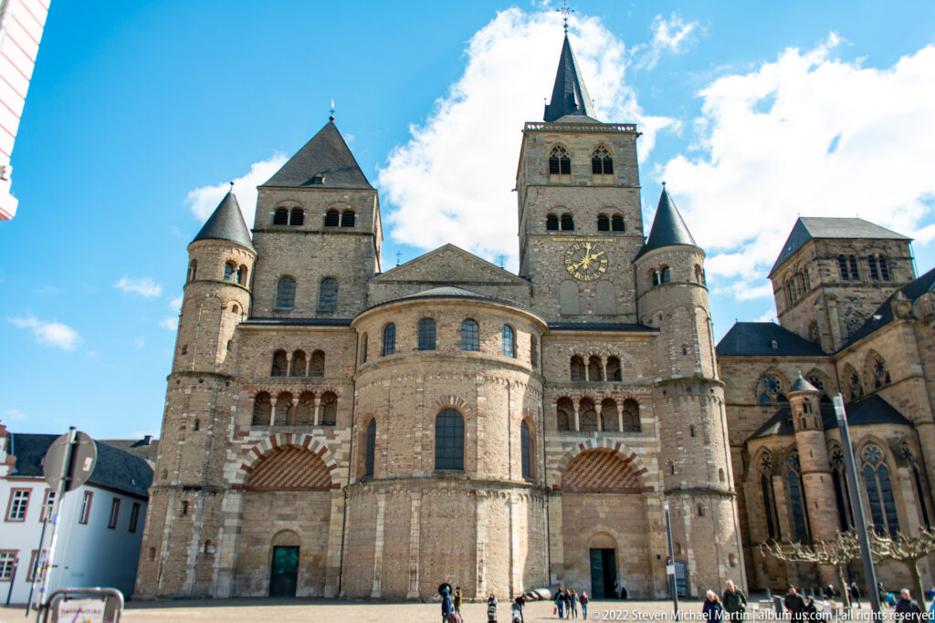 Massive stone exterior of Trier Cathedral with round apses, towers, arcades, and small visitors at the base.