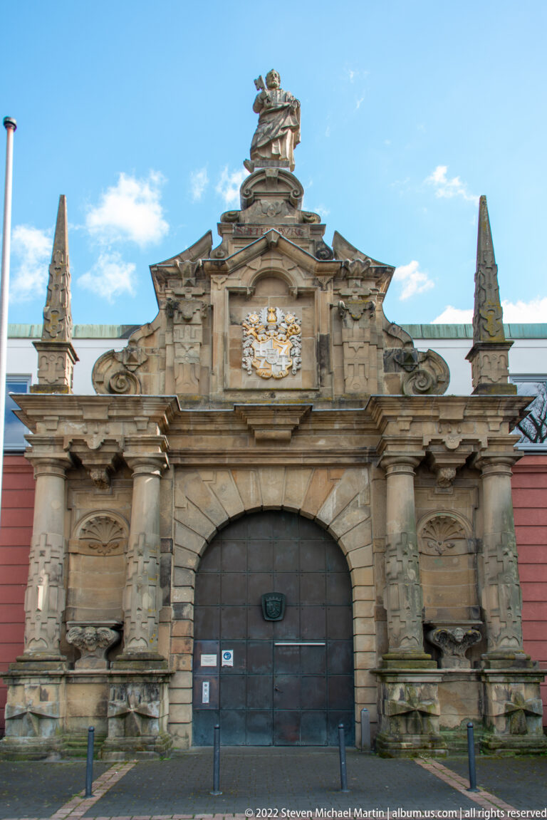 Large Baroque stone gateway with central arched doors, columns, sculpture, reliefs, and a pediment against a red-roofed wall.