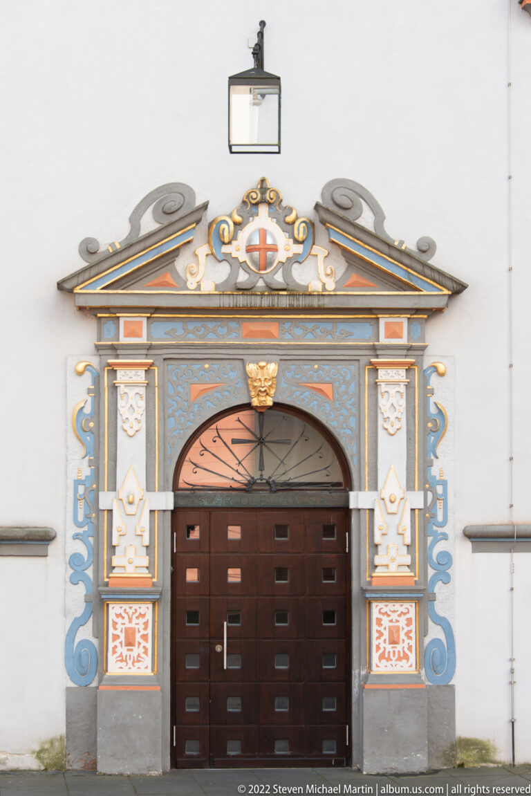 Dark wooden door framed by blue, white, orange, and gilded Baroque ornament beneath a triangular pediment.