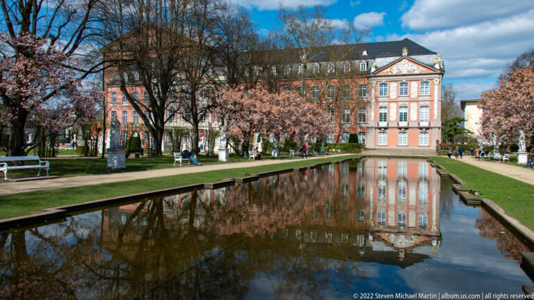 Electoral Palace in Trier reflected in a long formal garden pool bordered by lawns, trees, and paths.
