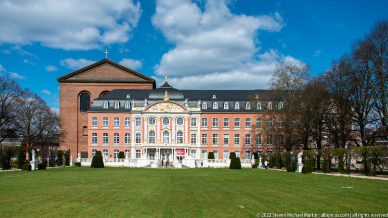 Long pink façade of Trier's Electoral Palace with ornate white-and-gold central pavilion seen across a formal lawn.