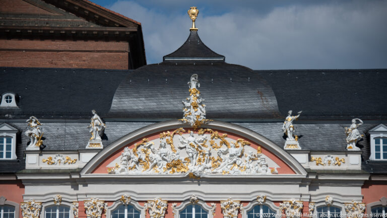 White-and-gold sculptural pediment with Rococo figures and scrollwork on Trier's Electoral Palace beneath dark slate roofs.